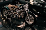 A custom-built motorcycle with polished metal components and intricate detailing, highlighted under soft lighting. The motorcycle features a shiny chrome engine, intricately designed exhaust pipes, and a light tan leather seat.