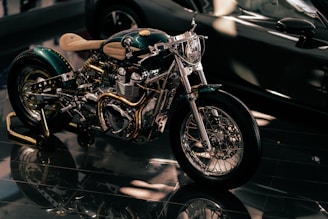 A custom-built motorcycle with polished metal components and intricate detailing, highlighted under soft lighting. The motorcycle features a shiny chrome engine, intricately designed exhaust pipes, and a light tan leather seat.