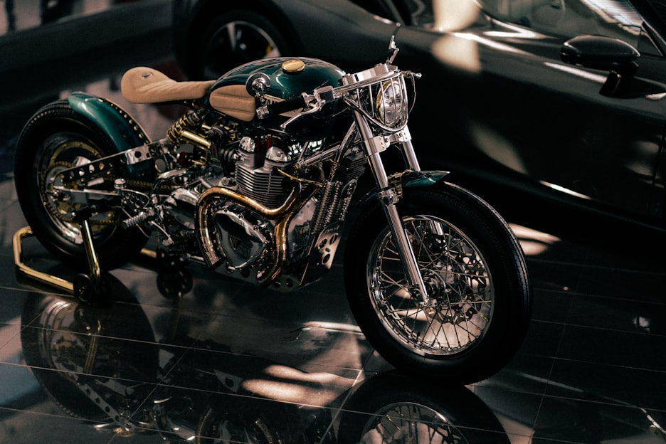 A custom-built motorcycle with polished metal components and intricate detailing, highlighted under soft lighting. The motorcycle features a shiny chrome engine, intricately designed exhaust pipes, and a light tan leather seat.