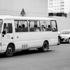 A white minibus on a city street with several passengers visible through the windows. The background includes a few parked cars and a building with a modern facade.