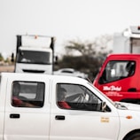 A white pickup truck is on the road with two occupants inside. The driver is focused and appears to be conversing with the passenger. In the background, a large white truck is visible, alongside a red delivery vehicle with text written on it. The setting is outdoors with some trees visible.