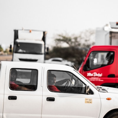 A white pickup truck is on the road with two occupants inside. The driver is focused and appears to be conversing with the passenger. In the background, a large white truck is visible, alongside a red delivery vehicle with text written on it. The setting is outdoors with some trees visible.
