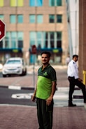 A man wearing a green and black uniform stands on a sidewalk. In the background, there is a security guard in a white shirt and a black tie, a stop sign, a white car, and a building with multiple windows.