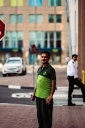 A man wearing a green and black uniform stands on a sidewalk. In the background, there is a security guard in a white shirt and a black tie, a stop sign, a white car, and a building with multiple windows.