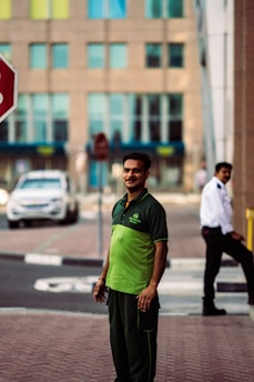 A man wearing a green and black uniform stands on a sidewalk. In the background, there is a security guard in a white shirt and a black tie, a stop sign, a white car, and a building with multiple windows.