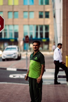 A man wearing a green and black uniform stands on a sidewalk. In the background, there is a security guard in a white shirt and a black tie, a stop sign, a white car, and a building with multiple windows.