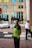 A man wearing a green and black uniform stands on a sidewalk. In the background, there is a security guard in a white shirt and a black tie, a stop sign, a white car, and a building with multiple windows.