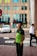 A man wearing a green and black uniform stands on a sidewalk. In the background, there is a security guard in a white shirt and a black tie, a stop sign, a white car, and a building with multiple windows.