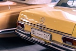 A vintage Mercedes-Benz car with a yellow exterior, featuring chrome details and a distinct rear design. The car has a Saudi Arabian license plate, and the lighting casts soft reflections on the surface.