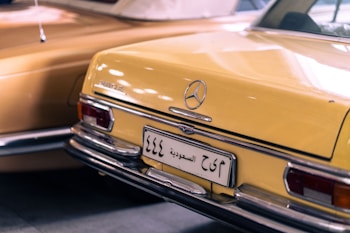 A vintage Mercedes-Benz car with a yellow exterior, featuring chrome details and a distinct rear design. The car has a Saudi Arabian license plate, and the lighting casts soft reflections on the surface.