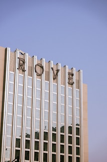 A tall building with a modern architectural style, featuring vertical glass panels and concrete sections. The top of the building displays large, stylized letters spelling 'ROME'. The sky above is clear and blue, creating a serene backdrop.