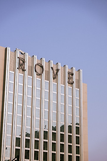 A tall building with a modern architectural style, featuring vertical glass panels and concrete sections. The top of the building displays large, stylized letters spelling 'ROME'. The sky above is clear and blue, creating a serene backdrop.