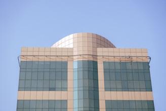 A modern building features a dome-shaped structure at the top, with a geometric design pattern. The façade is made up of rectangular glass panels, reflecting the azure sky, and is divided by horizontal and vertical lines.