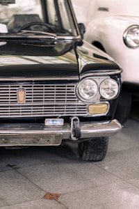 A vintage Fiat car is prominently featured, showcasing its front grille, headlights, and bumper. The vehicle is black and parked on a tiled floor, with part of an adjacent white car visible in the background. The environment suggests an indoor setting, possibly a showroom or museum.