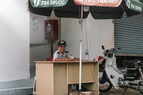 A person is seated at a small desk under a large umbrella. They are wearing a uniform with a cap and appear to be at a security post. To the side, there is a parked motorcycle next to the desk. A closed roll-up door is visible in the background, along with a red fire equipment box fixed on the wall.