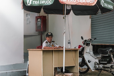A person is seated at a small desk under a large umbrella. They are wearing a uniform with a cap and appear to be at a security post. To the side, there is a parked motorcycle next to the desk. A closed roll-up door is visible in the background, along with a red fire equipment box fixed on the wall.