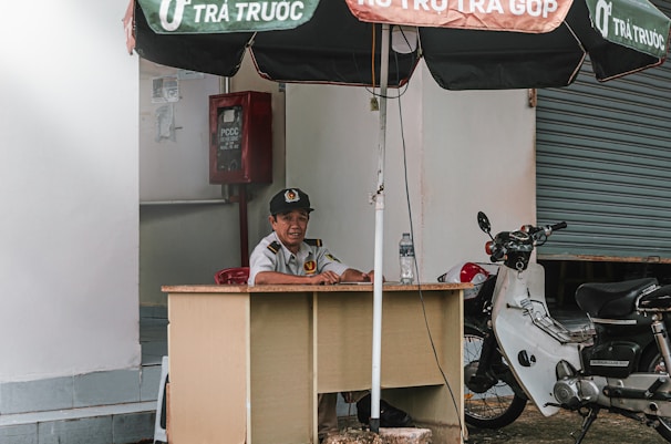 A person is seated at a small desk under a large umbrella. They are wearing a uniform with a cap and appear to be at a security post. To the side, there is a parked motorcycle next to the desk. A closed roll-up door is visible in the background, along with a red fire equipment box fixed on the wall.
