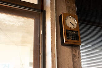 A wall-mounted, rectangular analog clock with a wooden frame and white clock face is displayed. It shows the time as almost 10 minutes past 10. Below the clock face, there is a digital calendar display showing the date. To the left of the clock, there is a window with partially visible stone or concrete, and to the right, there are closed venetian blinds.