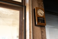 A wall-mounted, rectangular analog clock with a wooden frame and white clock face is displayed. It shows the time as almost 10 minutes past 10. Below the clock face, there is a digital calendar display showing the date. To the left of the clock, there is a window with partially visible stone or concrete, and to the right, there are closed venetian blinds.
