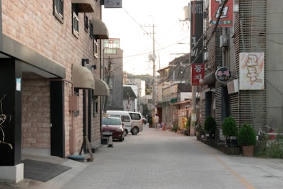 A narrow urban street lined with buildings on both sides, featuring signs with Korean text. The buildings showcase a mix of stone and concrete textures, with some potted plants placed by the entrances. The street is quiet, with a couple of parked vehicles including a white van and a red car. Overhead, power lines crisscross the sky, and a few pedestrians can be seen in the distance.