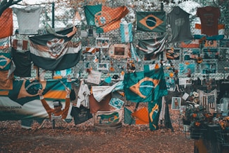 A memorial or tribute site featuring numerous flags, predominantly Brazilian, along with shirts, flowers, and photographs displayed on a chain-link fence. The ground is covered in fallen leaves, and there is a mix of vibrant and muted colors throughout.