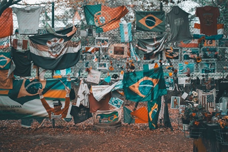 A memorial or tribute site featuring numerous flags, predominantly Brazilian, along with shirts, flowers, and photographs displayed on a chain-link fence. The ground is covered in fallen leaves, and there is a mix of vibrant and muted colors throughout.