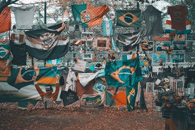 A memorial or tribute site featuring numerous flags, predominantly Brazilian, along with shirts, flowers, and photographs displayed on a chain-link fence. The ground is covered in fallen leaves, and there is a mix of vibrant and muted colors throughout.