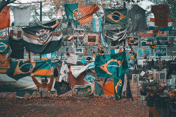 A memorial or tribute site featuring numerous flags, predominantly Brazilian, along with shirts, flowers, and photographs displayed on a chain-link fence. The ground is covered in fallen leaves, and there is a mix of vibrant and muted colors throughout.
