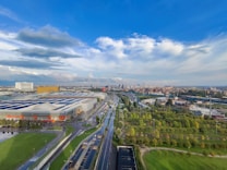 An expansive urban landscape featuring a large industrial building with solar panels on its roof. Roads and highways weave through the area, bordered by rows of lush green trees and landscaped parks. The cityscape in the distance includes various structures, and the sky is dominated by large clouds with patches of blue.