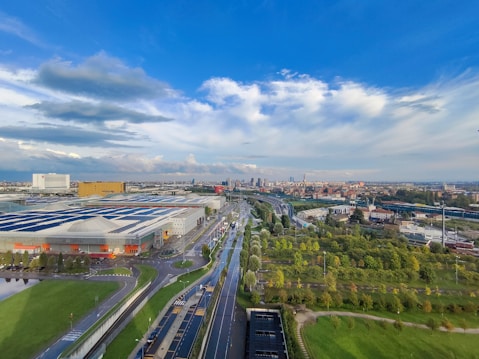 An expansive urban landscape featuring a large industrial building with solar panels on its roof. Roads and highways weave through the area, bordered by rows of lush green trees and landscaped parks. The cityscape in the distance includes various structures, and the sky is dominated by large clouds with patches of blue.