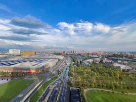 An expansive urban landscape featuring a large industrial building with solar panels on its roof. Roads and highways weave through the area, bordered by rows of lush green trees and landscaped parks. The cityscape in the distance includes various structures, and the sky is dominated by large clouds with patches of blue.