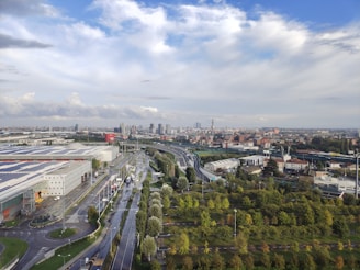 An urban landscape stretches into the distance with a mixture of industrial and residential buildings. A prominent highway runs through the cityscape, partially wet from recent rain. On one side, large warehouses with solar panels are visible, while lush green areas and lined trees decorate the foreground. The sky above is expansive with dramatic clouds providing a dynamic contrast against the blue.