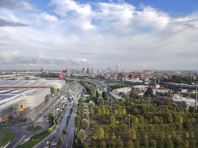 An urban landscape stretches into the distance with a mixture of industrial and residential buildings. A prominent highway runs through the cityscape, partially wet from recent rain. On one side, large warehouses with solar panels are visible, while lush green areas and lined trees decorate the foreground. The sky above is expansive with dramatic clouds providing a dynamic contrast against the blue.