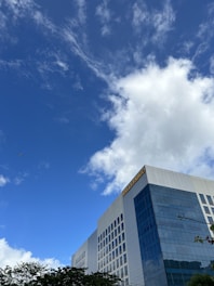 A modern, glass-fronted building with multiple floors stands tall against a backdrop of a bright blue sky with scattered clouds. The building has a prominent sign on the top corner. Trees with green leaves are visible at the bottom edge of the image.