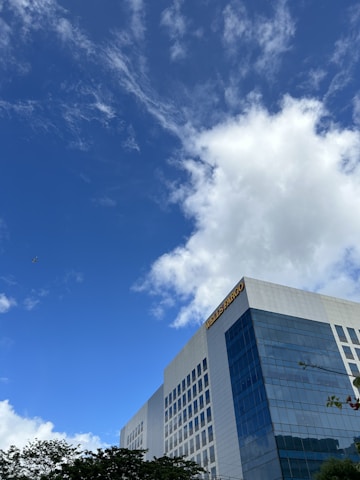 A modern, glass-fronted building with multiple floors stands tall against a backdrop of a bright blue sky with scattered clouds. The building has a prominent sign on the top corner. Trees with green leaves are visible at the bottom edge of the image.