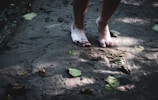 Bare feet standing on a sandy surface, partially covered in mud. Fallen leaves are scattered around on the ground, and shadows of foliage can be seen. The lighting suggests dappled sunlight filtering through trees.
