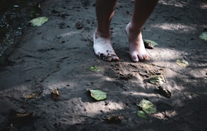 Bare feet standing on a sandy surface, partially covered in mud. Fallen leaves are scattered around on the ground, and shadows of foliage can be seen. The lighting suggests dappled sunlight filtering through trees.