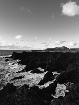 A dramatic black and white coastal scene featuring rocky cliffs and waves crashing against them. The sky is filled with scattered clouds, creating a contrast between the dark rocks and the bright sky.