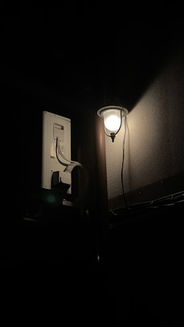 A wall-mounted lampshade illuminates a dark area, casting a soft glow on a textured wall. Nearby, electrical wiring and a device are attached to the wall, partially visible in the subdued light.