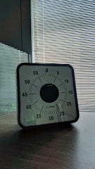 A mechanical timer with a black and white face featuring minutes marked in increments of five, set on a wooden table. The background consists of striped blinds with sunlight filtering through.