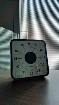 A mechanical timer with a black and white face featuring minutes marked in increments of five, set on a wooden table. The background consists of striped blinds with sunlight filtering through.