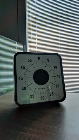 A mechanical timer with a black and white face featuring minutes marked in increments of five, set on a wooden table. The background consists of striped blinds with sunlight filtering through.