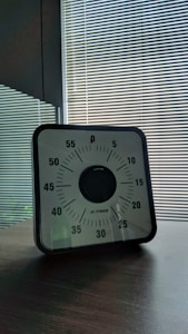 A mechanical timer with a black and white face featuring minutes marked in increments of five, set on a wooden table. The background consists of striped blinds with sunlight filtering through.