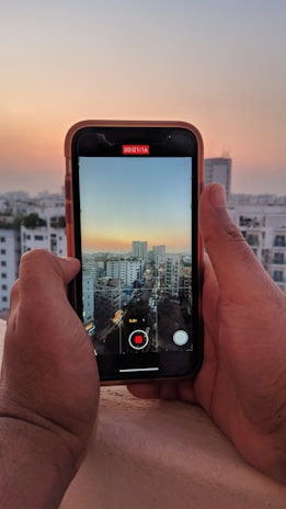 A person is holding a smartphone, capturing a video of a cityscape during sunset. The phone's screen shows the alignment with tall buildings under a gradient sky transitioning from blue to orange.