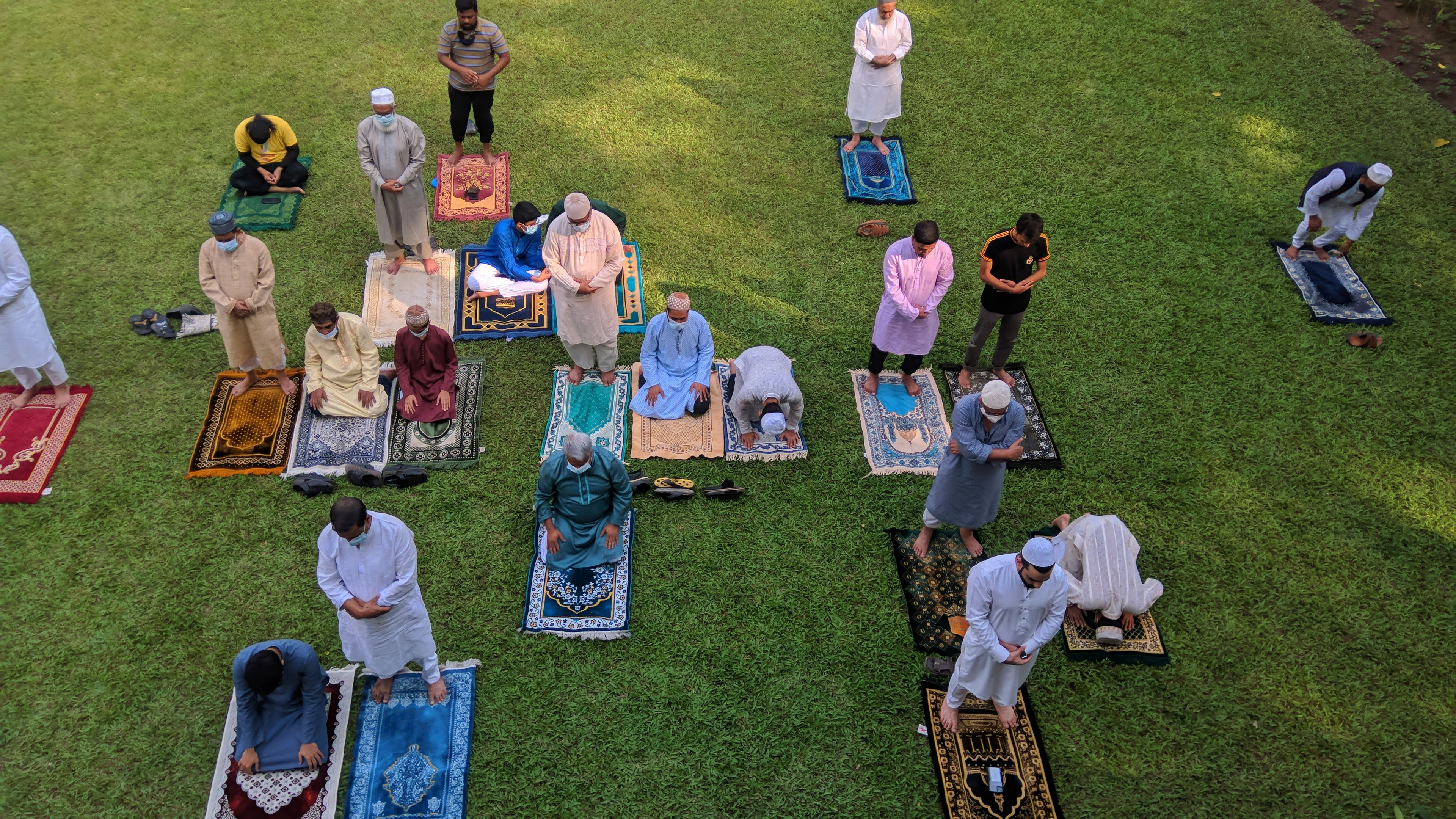 A group of people are gathered on a grassy area, each standing on colorful prayer rugs. They are positioned in rows, engaged in prayer. The scene appears serene and orderly, suggesting a communal activity or event.