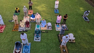 A group of people are gathered on a grassy area, each standing on colorful prayer rugs. They are positioned in rows, engaged in prayer. The scene appears serene and orderly, suggesting a communal activity or event.