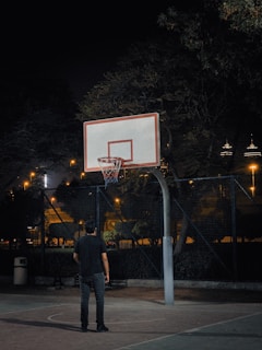 A nighttime scene featuring a person standing on an outdoor basketball court looking up at a basketball hoop. The court is surrounded by a chain-link fence and there are trees and city lights visible in the background.