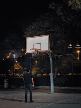 A nighttime scene featuring a person standing on an outdoor basketball court looking up at a basketball hoop. The court is surrounded by a chain-link fence and there are trees and city lights visible in the background.