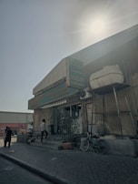 A small corner grocery store with signage in English and another language, likely Arabic. The exterior is worn and features a few potted plants. A bicycle is parked near the entrance, and a man in a checkered shirt is stepping inside. The sun is shining brightly from above, creating a high contrast with shadows. The surroundings include a paved pathway and a few people in the background.