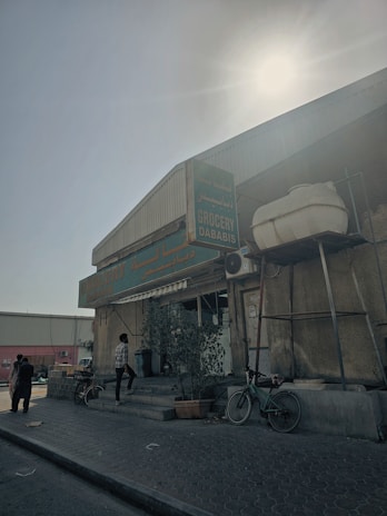 A small corner grocery store with signage in English and another language, likely Arabic. The exterior is worn and features a few potted plants. A bicycle is parked near the entrance, and a man in a checkered shirt is stepping inside. The sun is shining brightly from above, creating a high contrast with shadows. The surroundings include a paved pathway and a few people in the background.